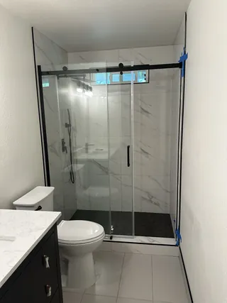 White veined shower wall tile beneath a high horizontal window, with black glass hardware, a dark shower floor and pale bathroom floor tile.