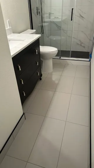 Pale rectangular bathroom floor tile beside a dark vanity and glass shower enclosure.