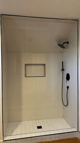 Front view of a white tiled shower with a horizontal niche, black overhead and handheld fittings, slim rectangular floor mosaic and a square drain opening.