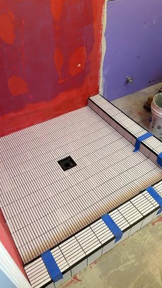 Slim rectangular mosaic tile on a shower floor and threshold beside red-coated wall areas, with blue tape and a square drain opening.