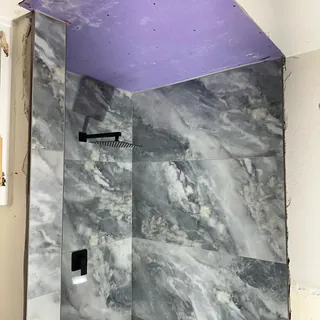Square black overhead showerhead and control against cloudy gray-veined wall tile beneath an unfinished purple ceiling.
