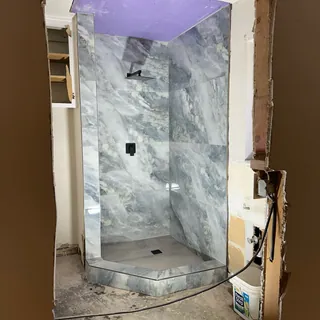 Corner shower with cloudy gray-veined rectangular tile, black overhead head and control, square drain and angled threshold.