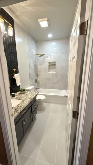 Updated bathroom with a gray vanity, dark rectangular accent tile, pale veined tub-surround tile, a divided niche and warm metal fittings.