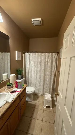 Brown vanity and beige square floor tile beside a toilet and curtained tub in the original bathroom.