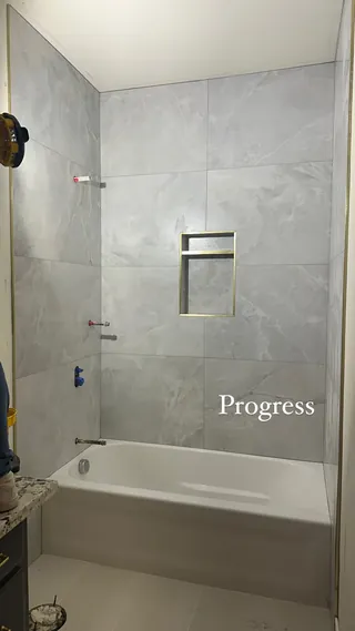 Pale gray rectangular veined tile around a white tub, with a gold-tone two-compartment recessed niche and unfinished fitting openings.