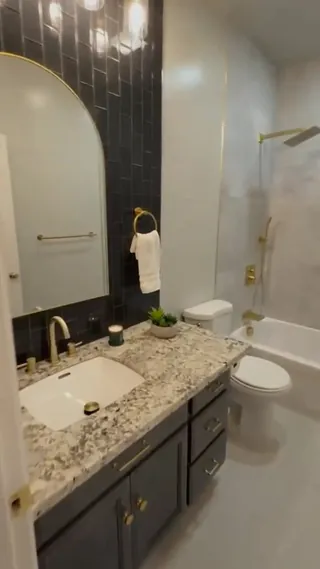 Gray vanity with a mottled light countertop, arched mirror and dark rectangular accent tile beside a white tub with warm metal fittings.