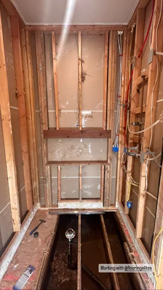 Exposed framing and an open floor cavity in a narrow shower area, with visible fixture pipes on the right.