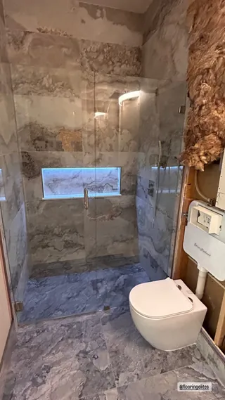 Veined rectangular shower wall and floor tile, a blue-lit horizontal niche and clear glass beside a wall-hung toilet; exposed framing remains to the right.