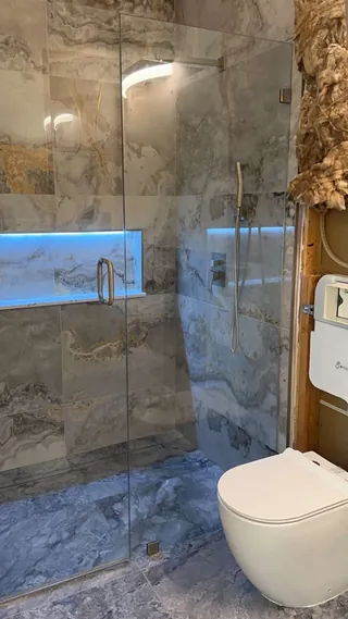 Blue-lit horizontal niche in veined rectangular wall tile behind glass, with a handheld shower and wall-hung toilet beside exposed insulation.