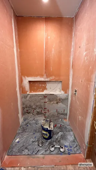Veined rectangular tile being installed across the shower floor and lower rear wall, with coated wall areas, spacers and tools visible.