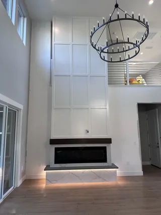 Tall white paneled wall above a pale veined fireplace surround, dark mantel and illuminated hearth.