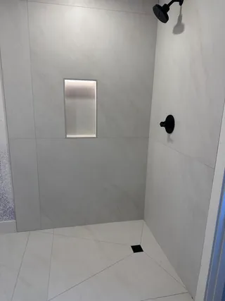 Pale veined shower tile around a tall illuminated niche, with black shower fittings, a square dark drain and diagonal floor cuts.