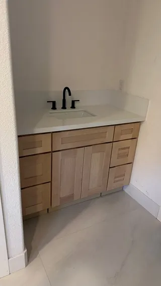 Light wood-tone vanity below a white basin, with pale floor tile, a bare wall above and an open doorway beside it.