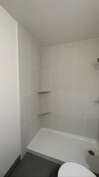 Two triangular shelves sit in the corner of a pale tiled shower above a low white base.
