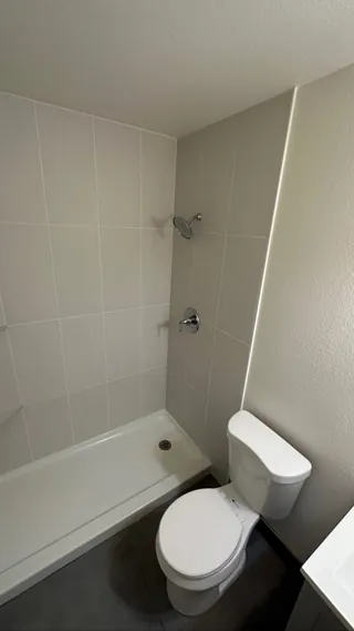 Low white shower base with a round drain beside gray floor tile and a toilet.