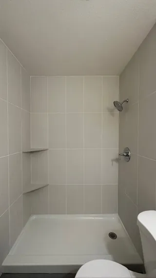 Pale rectangular wall tiles surround a low white shower base with two triangular corner shelves and chrome fittings.