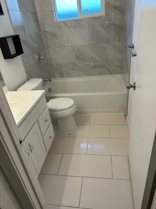 Gray-veined rectangular tile surrounds a white bathtub beneath a horizontal window, beside a white vanity, toilet and pale offset floor tile.