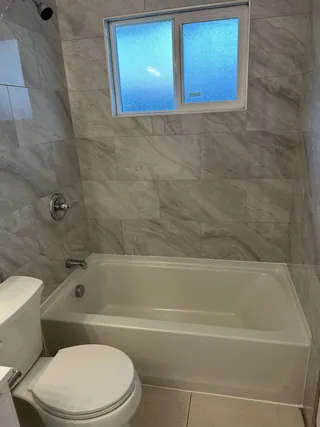 White bathtub beneath a horizontal window, with gray-veined rectangular wall tile and silver-tone shower head, control and tub spout.