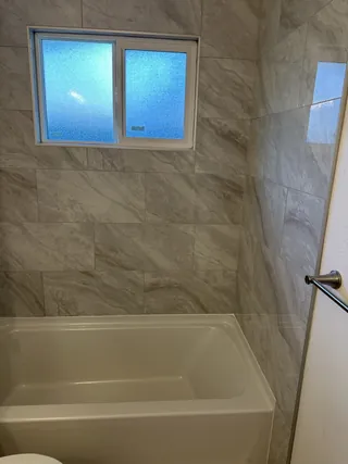Gray-veined rectangular tile around a horizontal sliding window above a white bathtub, viewed from the opposite side of the alcove.