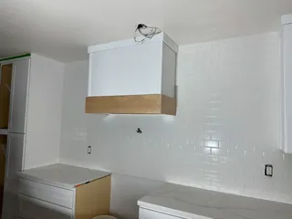 White running-bond subway tile rises around a box hood with a wood-tone band between white cabinets.