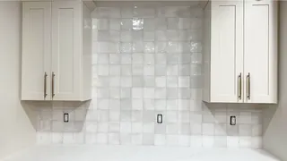 Glossy pale square backsplash tiles form a straight grid between white cabinets with warm-metal handles.