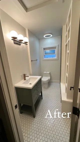 Narrow bathroom with white hexagonal mosaic floor tile, green-gray vanity, open shelving and a window at the far end.