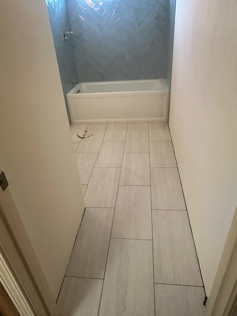 Rectangular floor tile leading toward a white bathtub with blue-gray herringbone wall tile beneath a horizontal window.