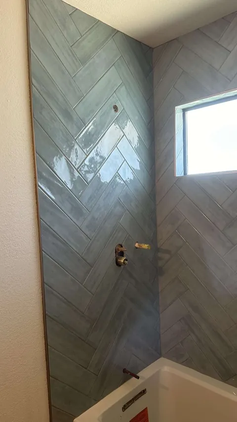 Blue-gray herringbone tile on the left wall of a tub alcove, with unfinished fixture openings and a horizontal window.