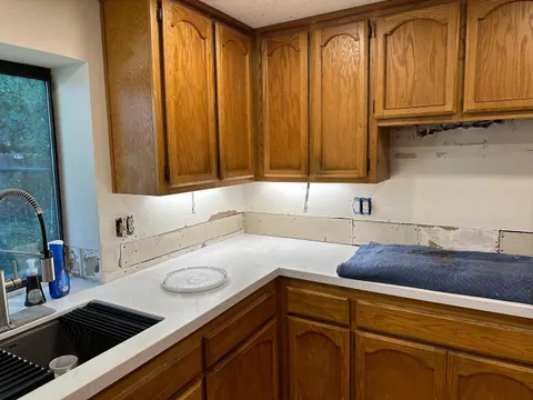 Bare backsplash wall beneath wood cabinets at a kitchen inside corner, with protected counters and exposed outlets.