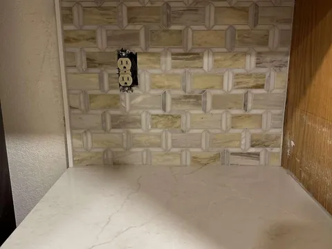 Rectangular mosaic backsplash above a narrow pale counter between a wall and wood cabinet, with an uncovered outlet.