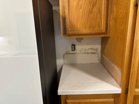 Bare backsplash wall above a narrow pale counter between a refrigerator and wood cabinet, with an exposed outlet.