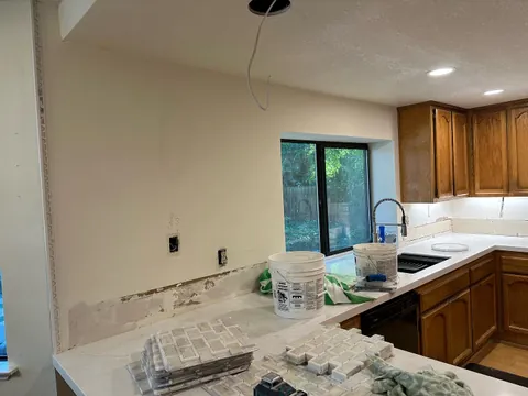 Kitchen wall around a window before backsplash tile, with exposed outlets, tile sheets and work supplies on the counter.