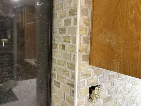 Close view of rectangular mosaic tile and pale edge trim on the right side of a dark kitchen window, with an uncovered outlet below a wood cabinet.