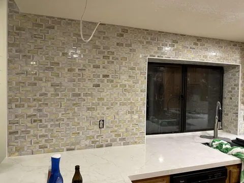 Cream and gray rectangular mosaic backsplash around a dark kitchen window above a pale countertop.