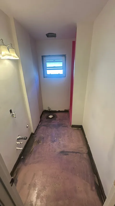 Stripped narrow bathroom with exposed floor, plumbing openings and a window at the far end.