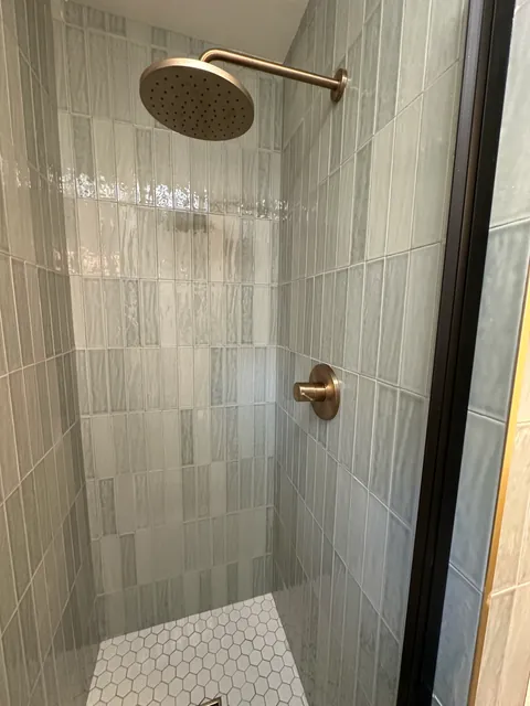 Round warm-metal shower head against vertical gray-green rectangular wall tile.