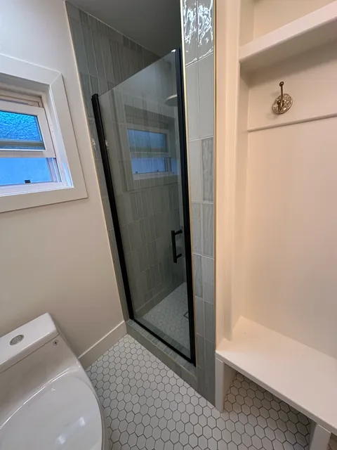 Glass shower enclosure beside the window and open shelves, with white hexagonal floor tile.