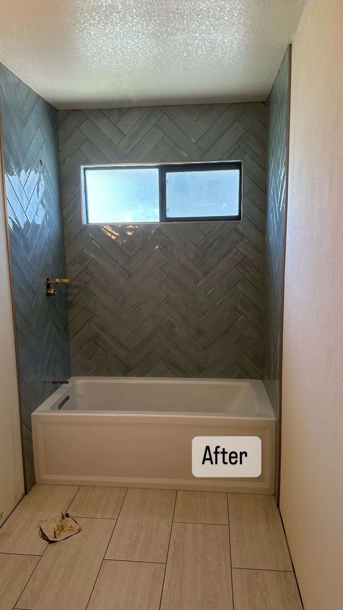 Blue-gray herringbone tile surrounding a white bathtub and horizontal window, with rectangular tile on the floor.