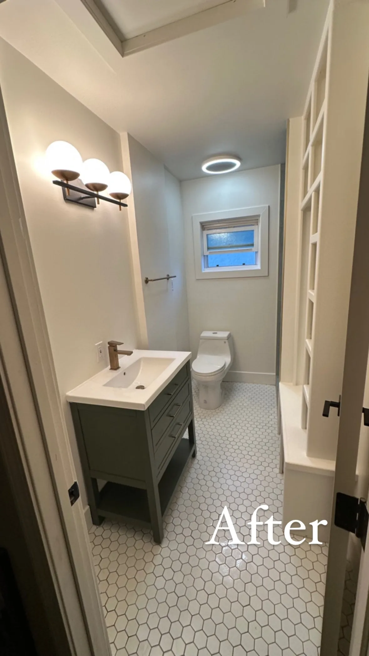 Narrow bathroom with white hexagonal mosaic floor tile, green-gray vanity, open shelving and a window at the far end.