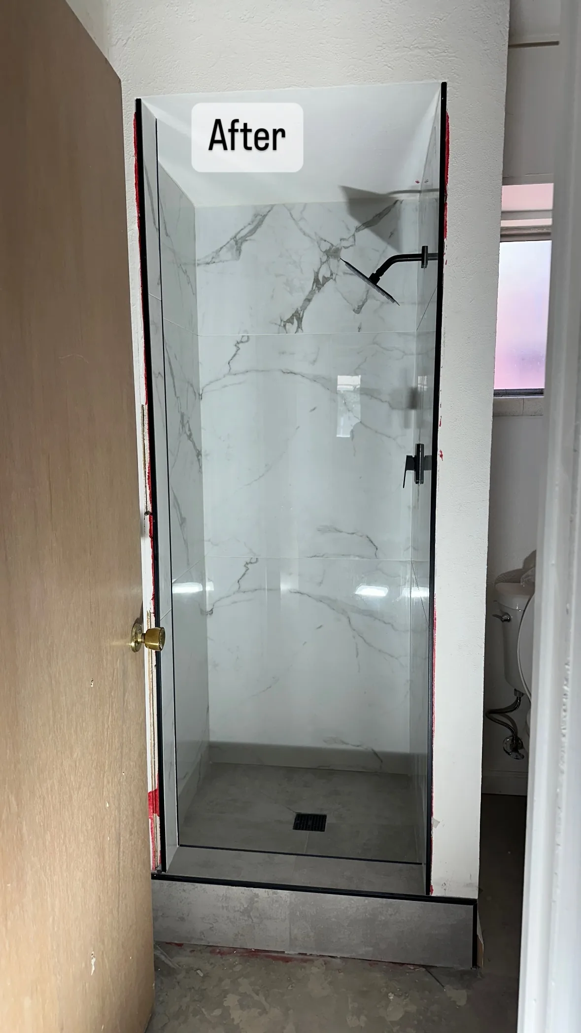 White veined shower wall tile behind clear glass, with a black shower head, tiled threshold and square drain.